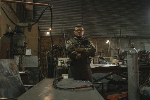 Free A bearded blacksmith stands confidently in an industrial workshop surrounded by tools. Stock Photo