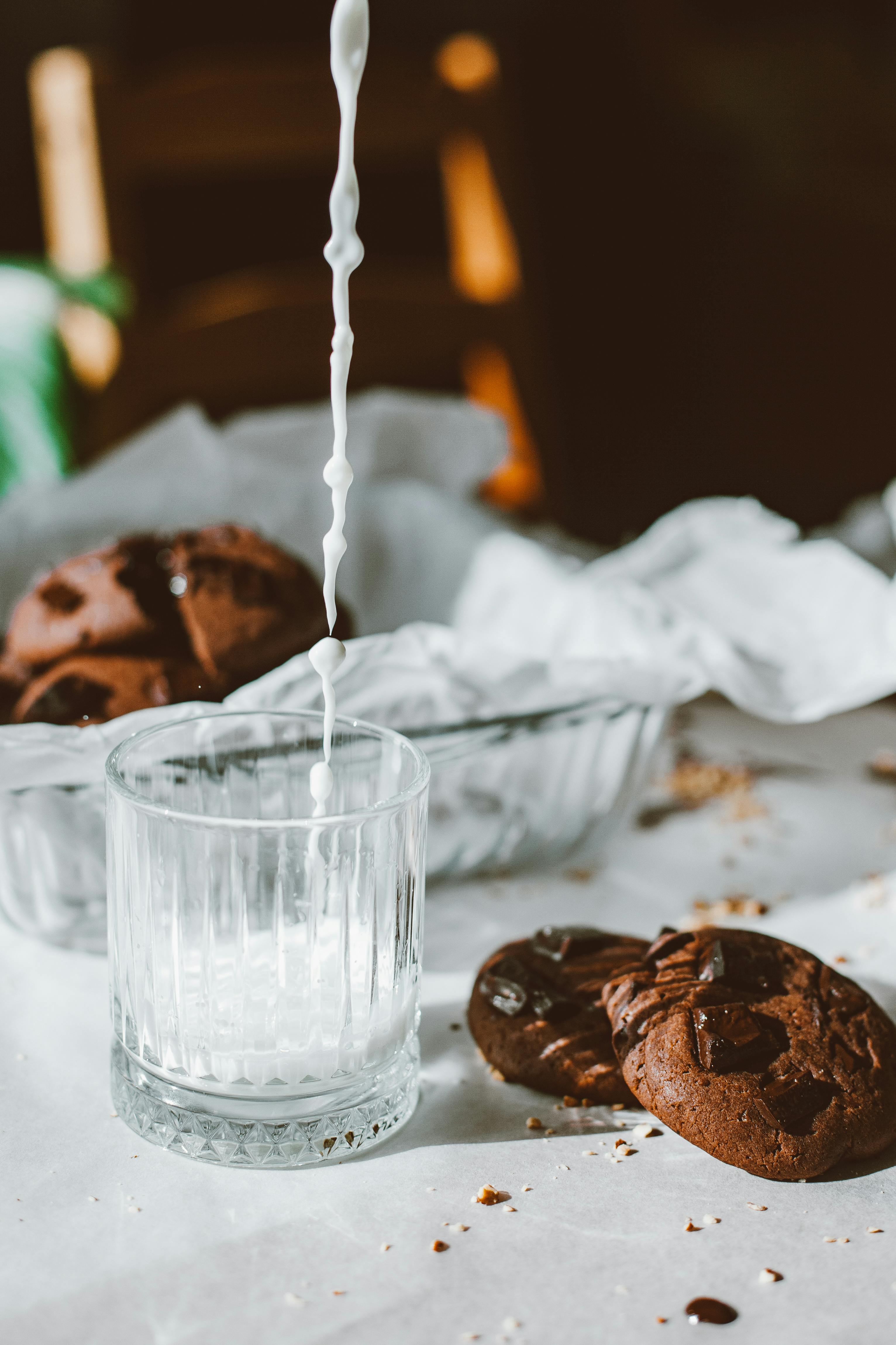 A close-up of chocolate cookies and milk pouring into a glass on a sunny kitchen counter.
