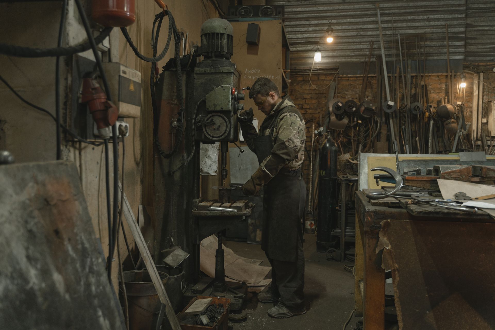 Bearded blacksmith using a large drill in an industrial workshop setting with various tools and equipment.