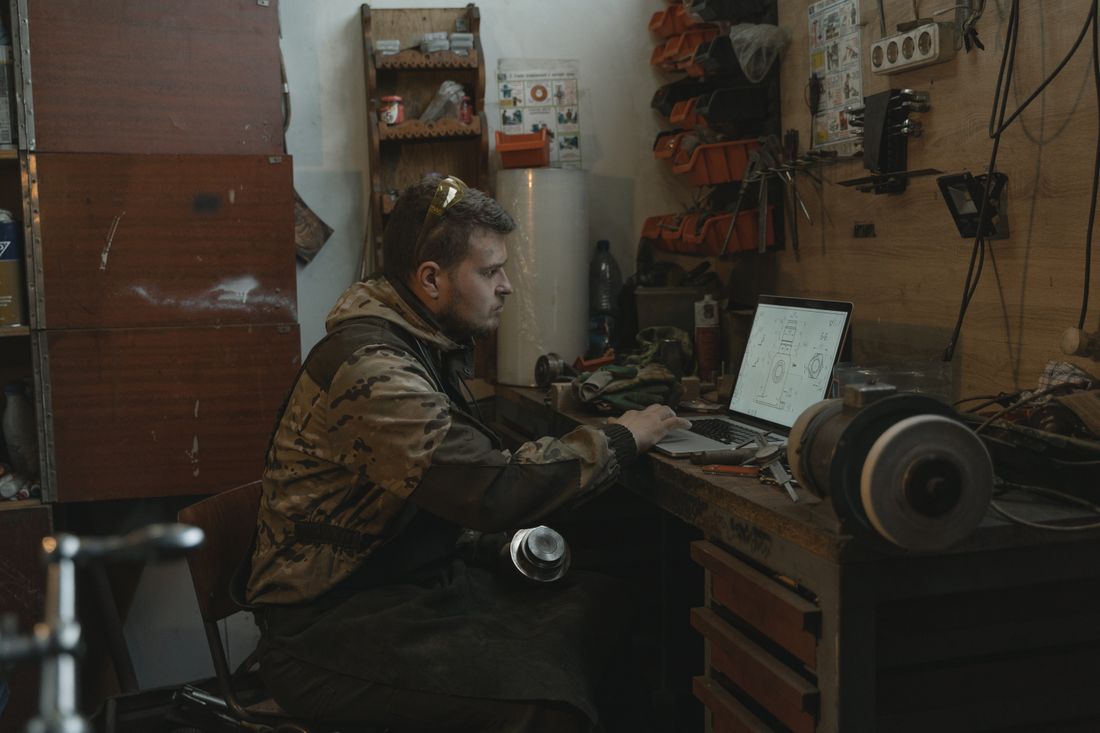 Mężczyzna w kurtce roboczej przy stole warsztatowym ogląda rysunek techniczny na laptopie