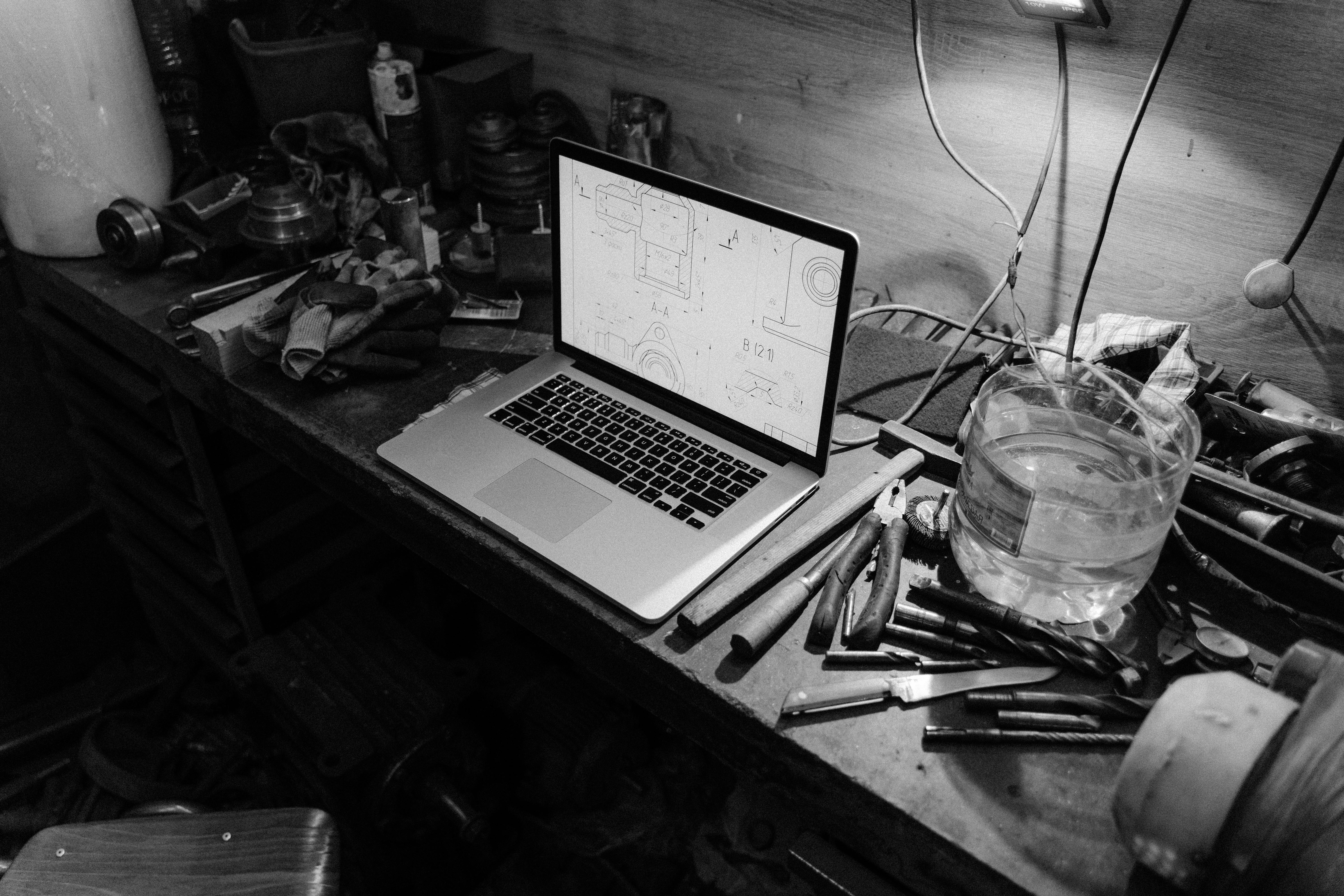 Black and white image of a workshop with a laptop displaying blueprints on a workbench, surrounded by tools.