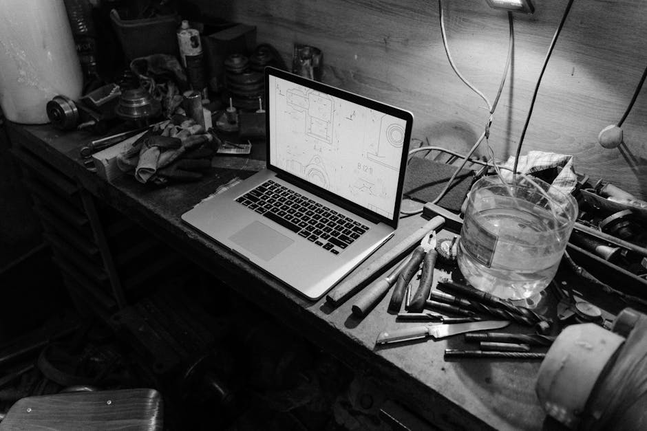 Black and white image of a workshop with a laptop displaying blueprints on a workbench, surrounded by tools.