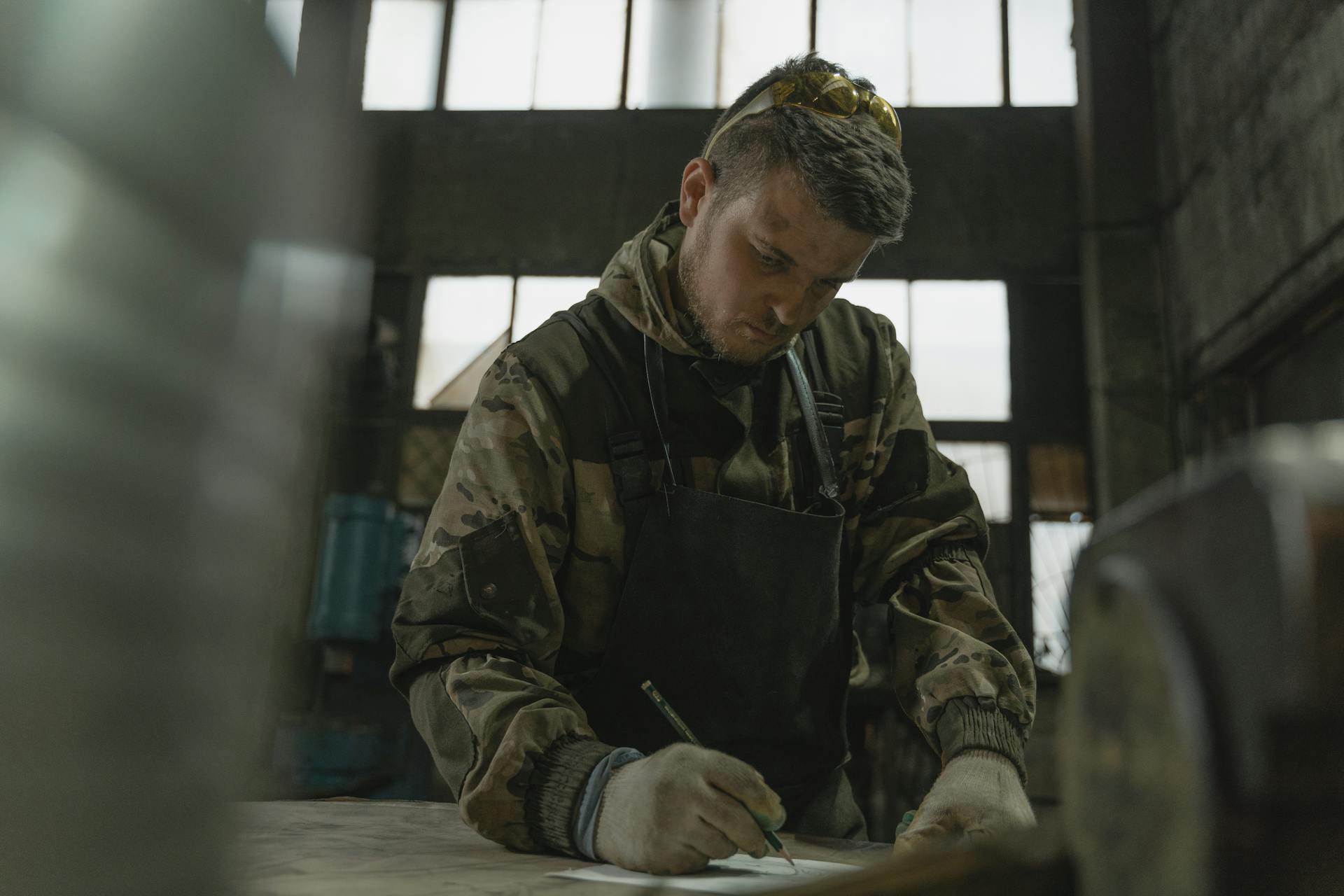 A man sketches on paper in a dimly lit industrial factory setting, wearing camouflage attire.