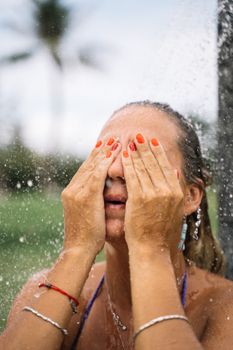 Woman enjoying a refreshing outdoor shower, covering her face with hands under clear summer sky.