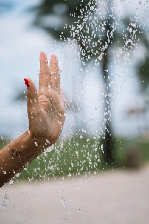 Free A hand with red painted nails touches falling water droplets, creating a refreshing outdoor feel. Stock Photo