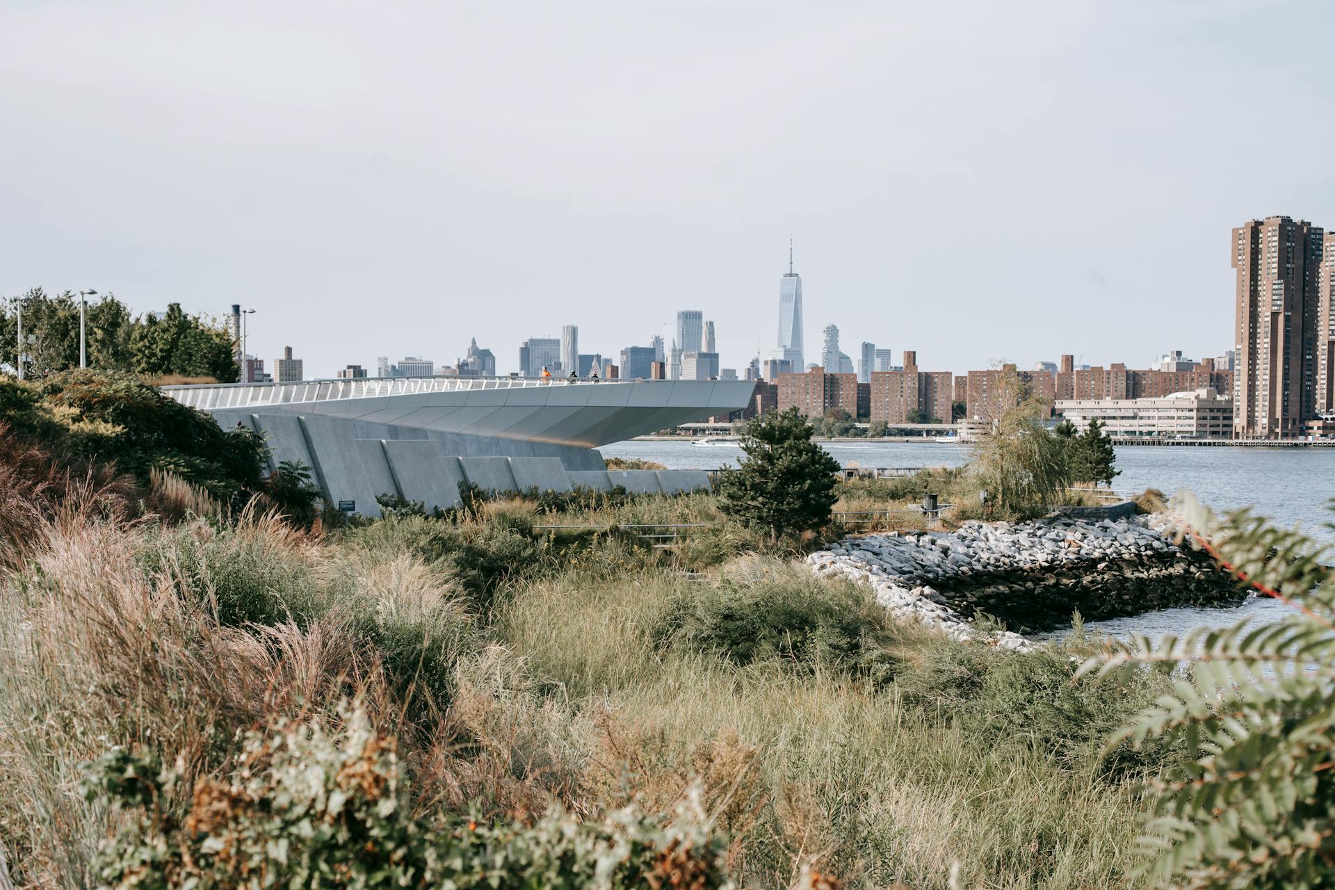 Tranquil Hudson River flowing between grassy embankment and modern city district with high rise skyscrapers and residential buildings placed in New York City in daytime