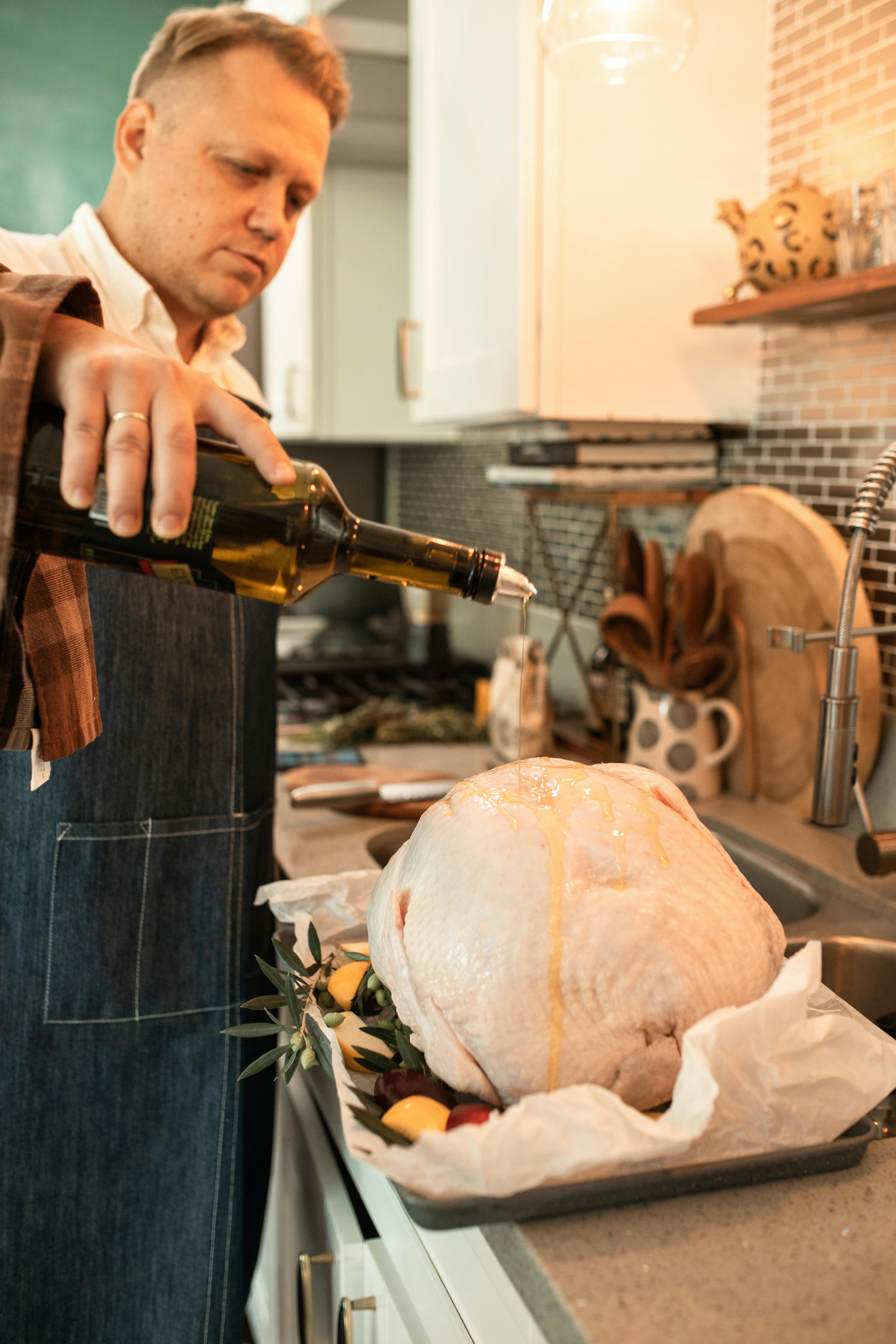 A man in a kitchen pouring oil over a raw turkey, setting the stage for a Thanksgiving feast.
