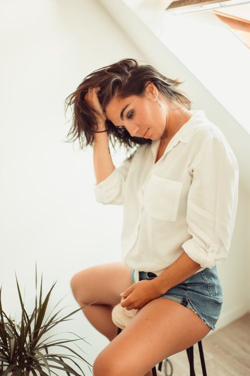 Free A relaxed woman in a white shirt and denim shorts indoors, near a plant. Stock Photo
