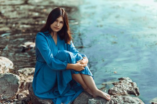 Free A serene portrait of a woman in a blue dress sitting by the lakeside, embracing a calm and picturesque setting. Stock Photo