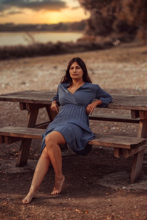 Free A woman in a blue dress sits barefoot on a bench at a serene beach during sunset. Stock Photo