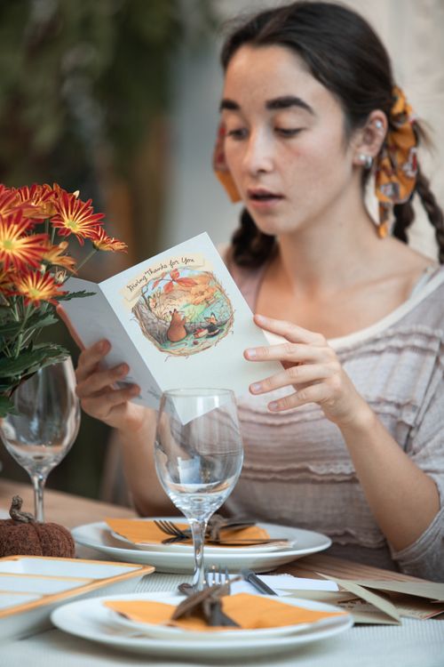 Free Young woman reading a card at a Thanksgiving dinner table, indoors with autumn decor. Stock Photo