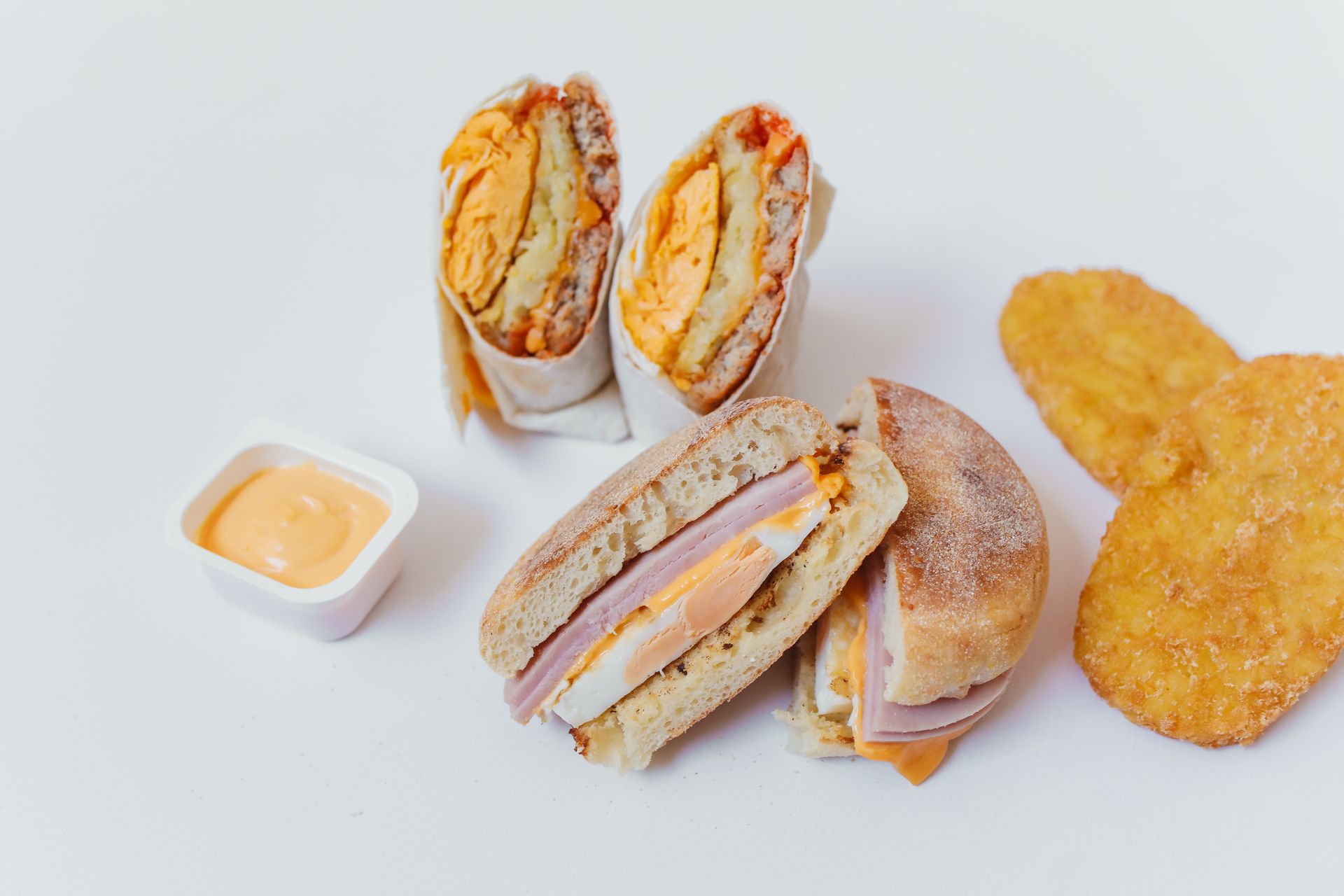A mouth-watering breakfast spread featuring a sandwich, wrap, and hash browns on a white surface.