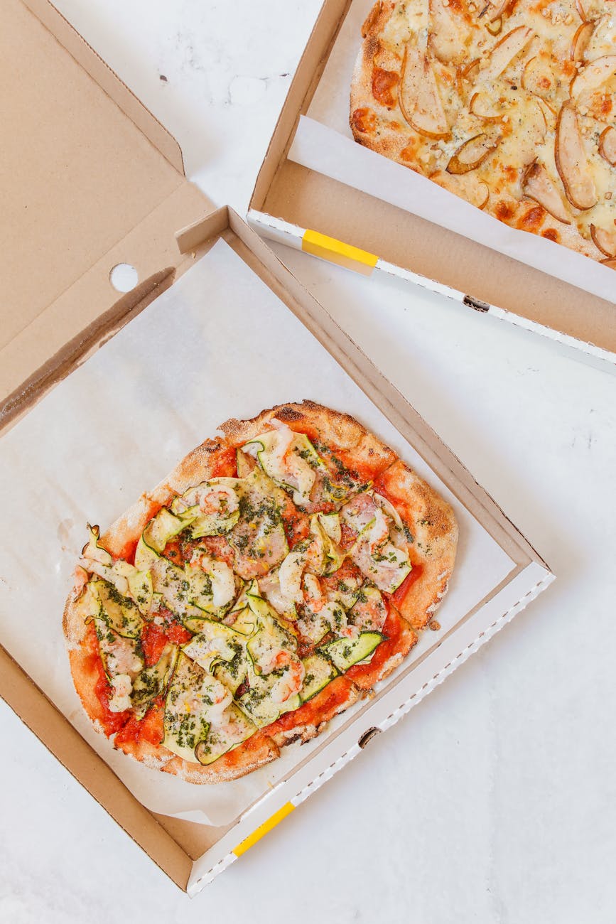 Top view of Italian pizzas in cardboard boxes with zucchini and pear toppings.