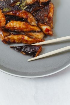用筷子夹起一道美味的中国茄子菜肴，在灰色盘子上进行特写拍摄，充分展现其诱人的亚洲风味