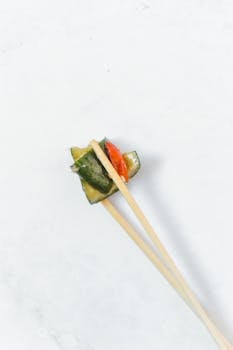 Overhead shot of cucumbers held by chopsticks, against a clean white surface.