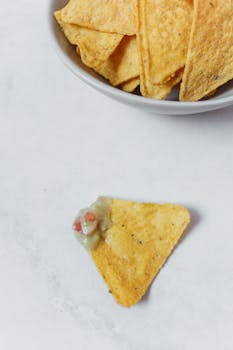 A bowl of tortilla chips with a single nacho topped with guacamole on a light background.