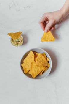 A hand holding a nacho over a bowl with dipping sauce, perfect for snack lovers.