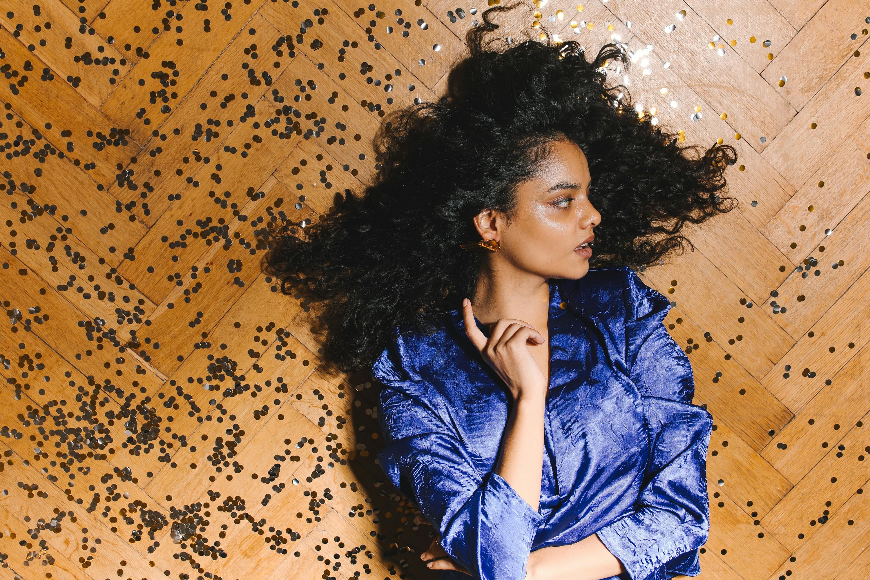 一名卷发女子身着蓝色连衣裙，在洒满五彩纸屑的木地板上摆出姿势