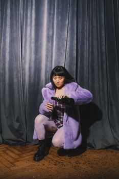 一位身穿紫色外套的亚洲妇女在窗帘背景下将葡萄酒倒入玻璃杯