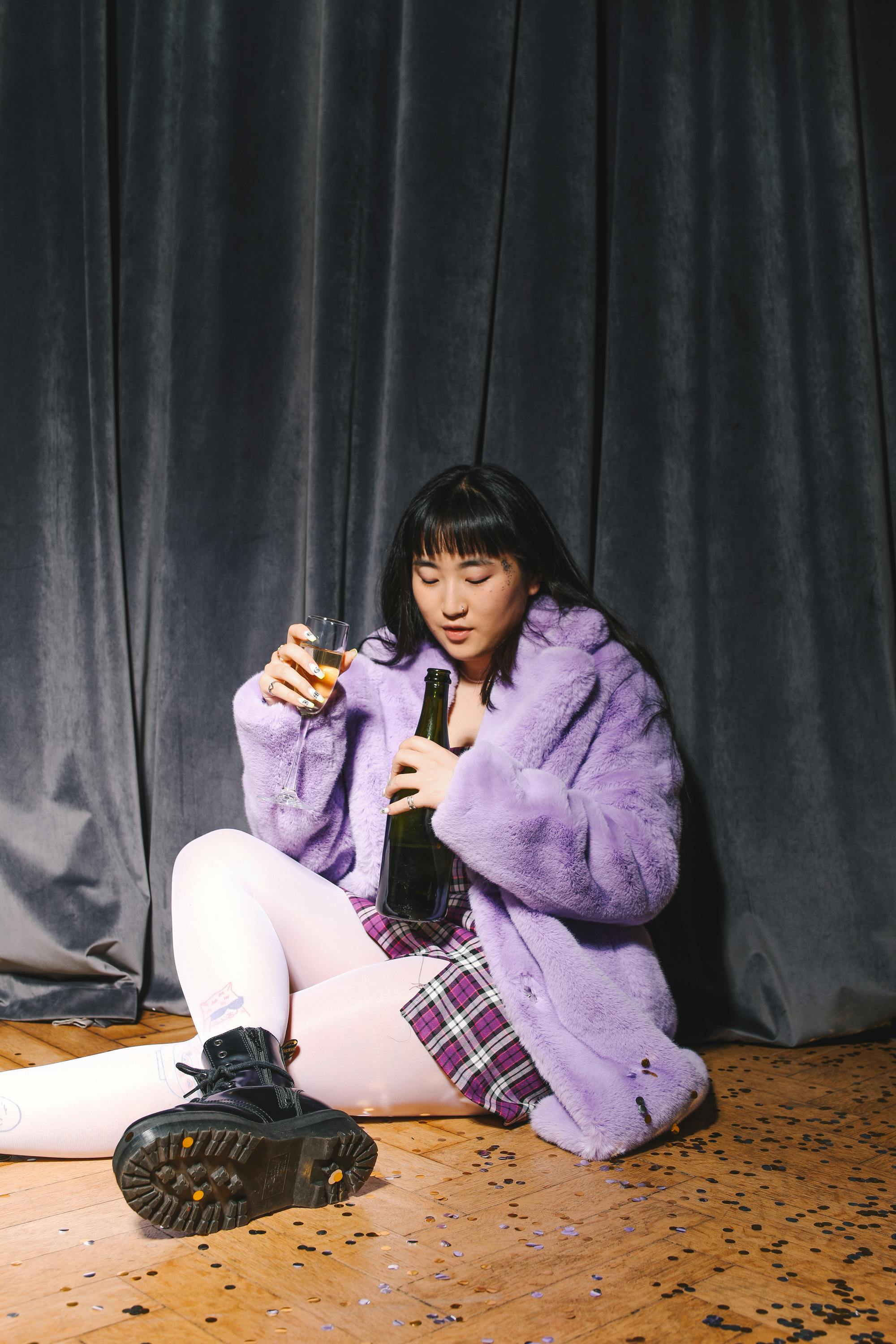一位穿着紫色外套的年轻女子坐在室内，手里拿着酒瓶和玻璃杯