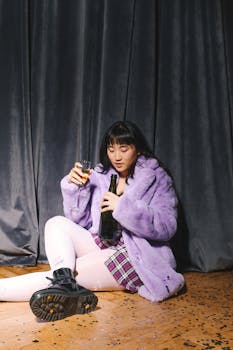 一位穿着紫色外套的年轻女子坐在室内，手里拿着酒瓶和玻璃杯