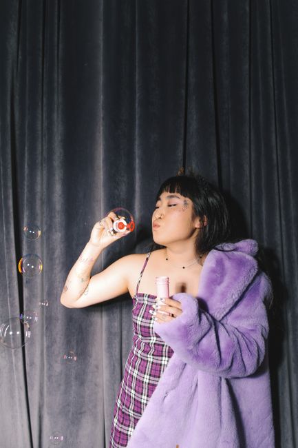 Fashionable woman in a purple coat blowing bubbles against a dark curtain backdrop.