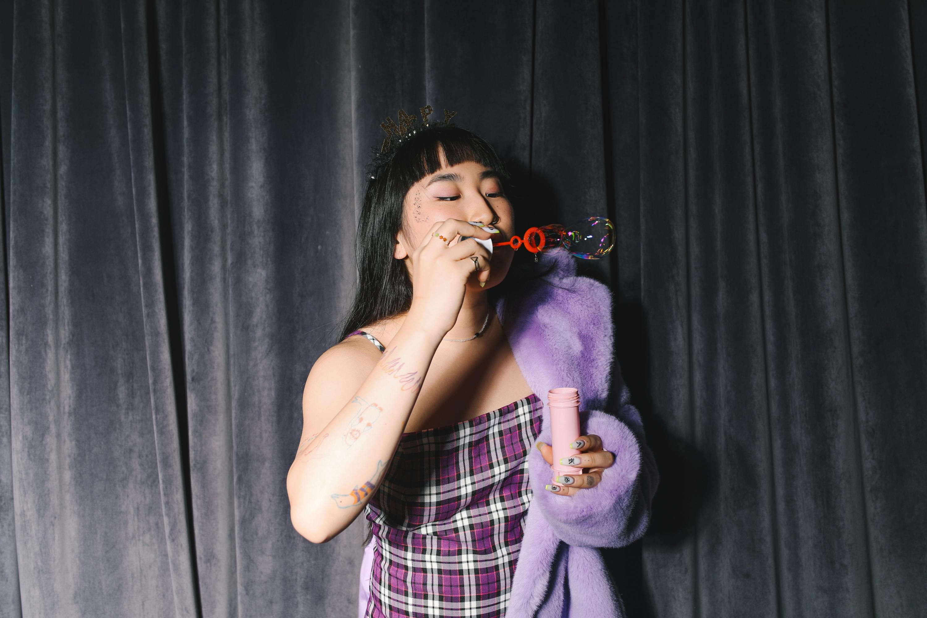 A woman wearing a plaid dress and purple coat blows bubbles indoors against a curtain.