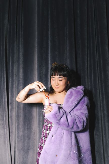 Asian woman wearing a purple fur coat blowing bubbles against a dark background.
