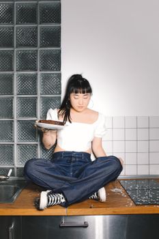 在现代厨房里，一名年轻女子盘腿坐在台面上，手里拿着一块巧克力蛋糕