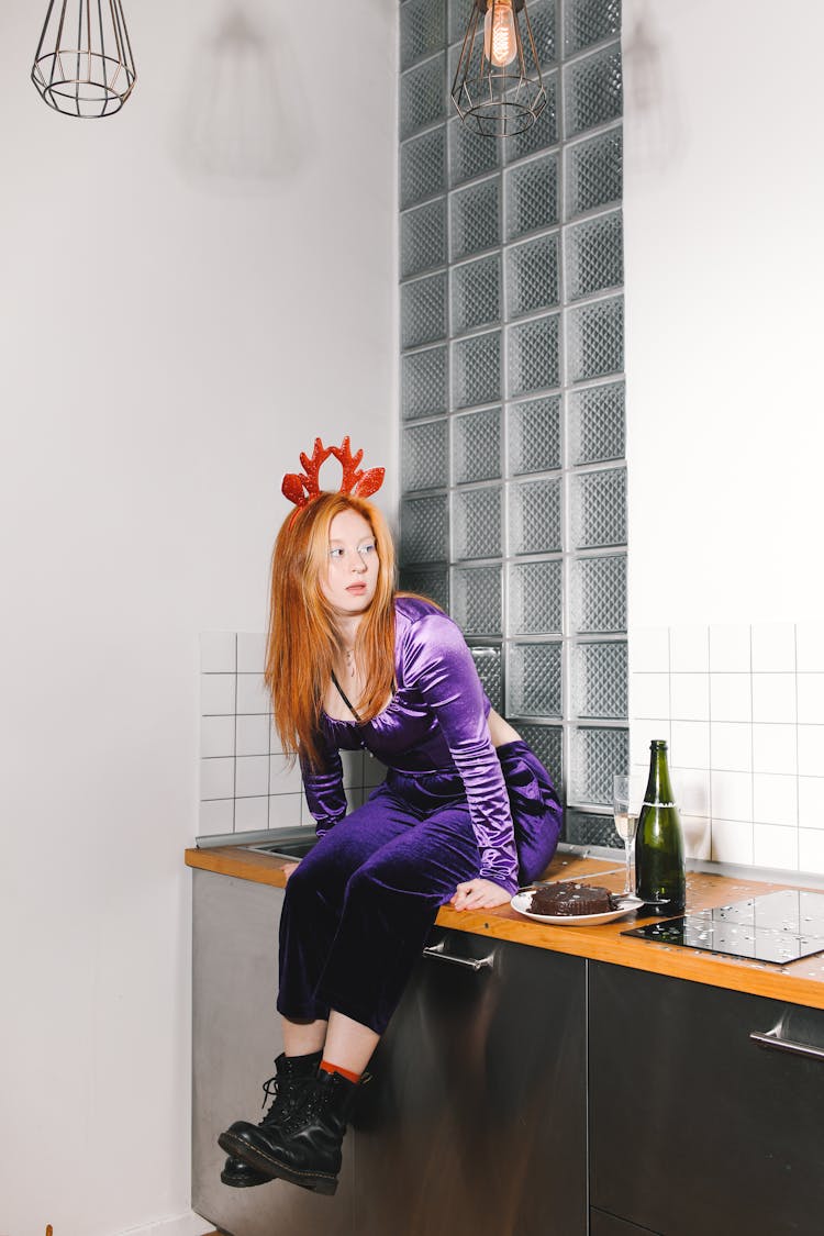 Young Woman Wearing A Reindeer Antlers Headband Sitting On The Kitchen Counter With Cake And Bottle Of Champagne​