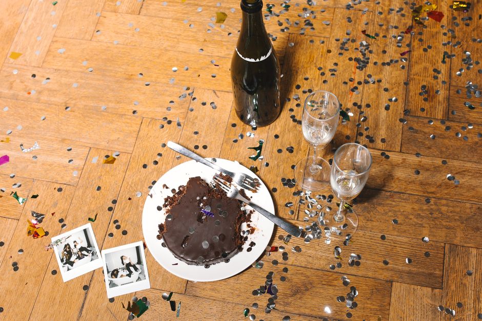 Post-party scene with a chocolate cake, champagne bottle, and confetti on a wooden floor.