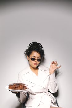一位时尚女士在室内穿着白外套、戴着太阳镜，拿着蛋糕摆姿势庆祝特殊时刻