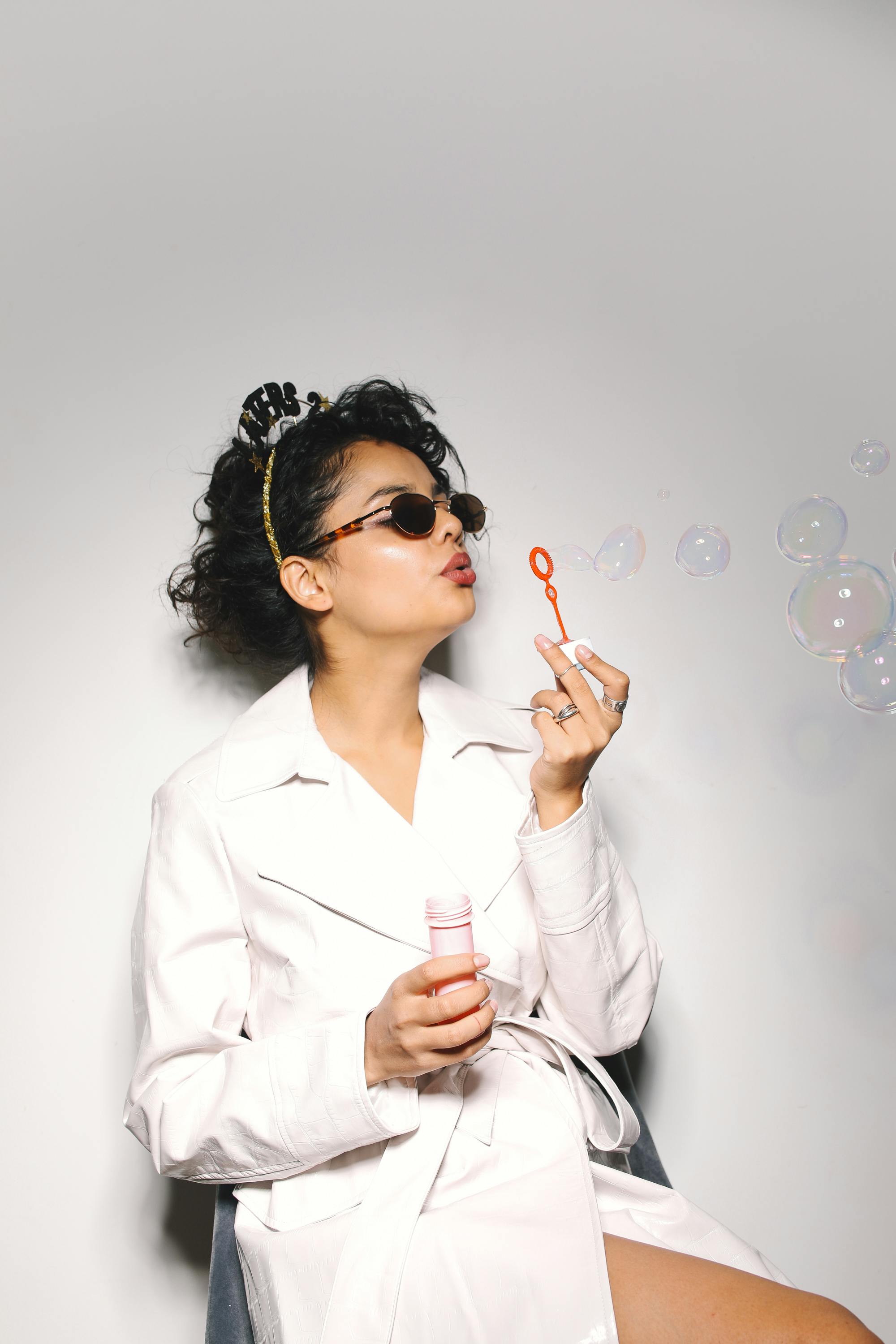 Fashionable woman in sunglasses blowing bubbles indoors, wearing a trench coat.