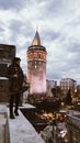 Full length contemplative bearded ethic male in warm clothes standing on snowy rooftop against medieval stone Galata Tower in Istanbul on cloudy winter day