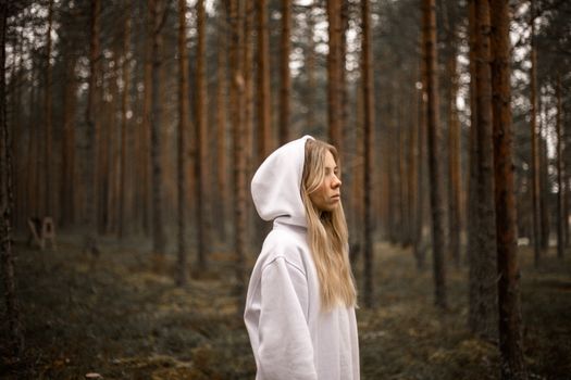 一位身着白色连帽衫的年轻女子，正静静伫立于森林之中，陷入沉思