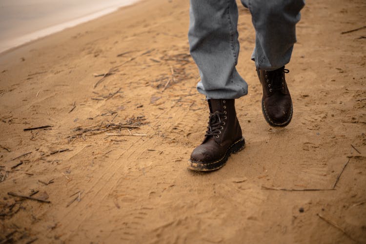 Person In Blue Denim Jeans And Black Leather Boots Walking On Brown Sand