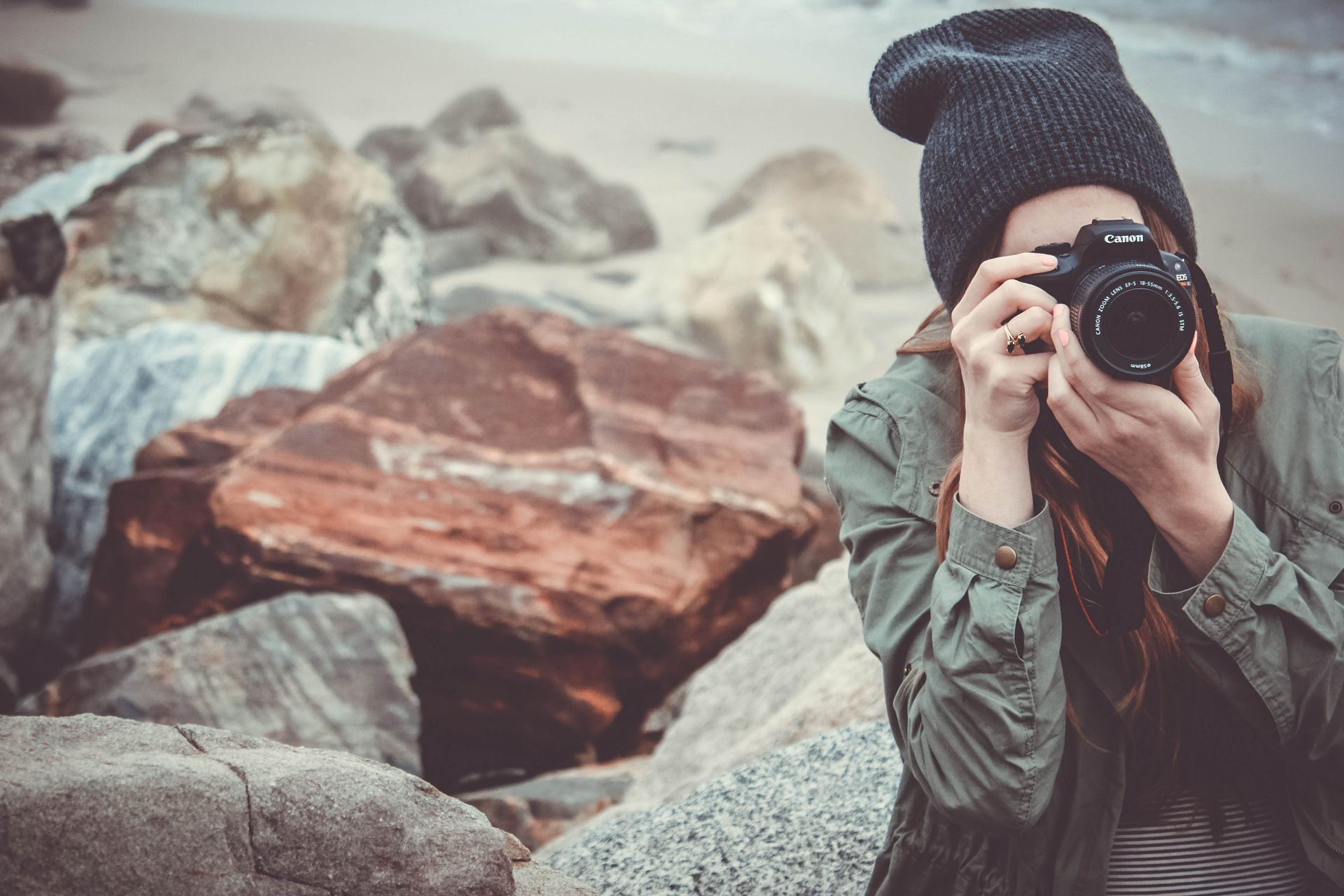 Woman taking a photograph on a rocky beach with a Canon DSLR camera.