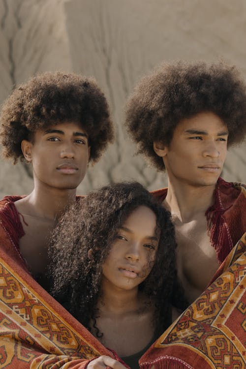 Free Three young individuals with afro hair wrapped in printed cloth, standing in a sandy desert environment. Stock Photo
