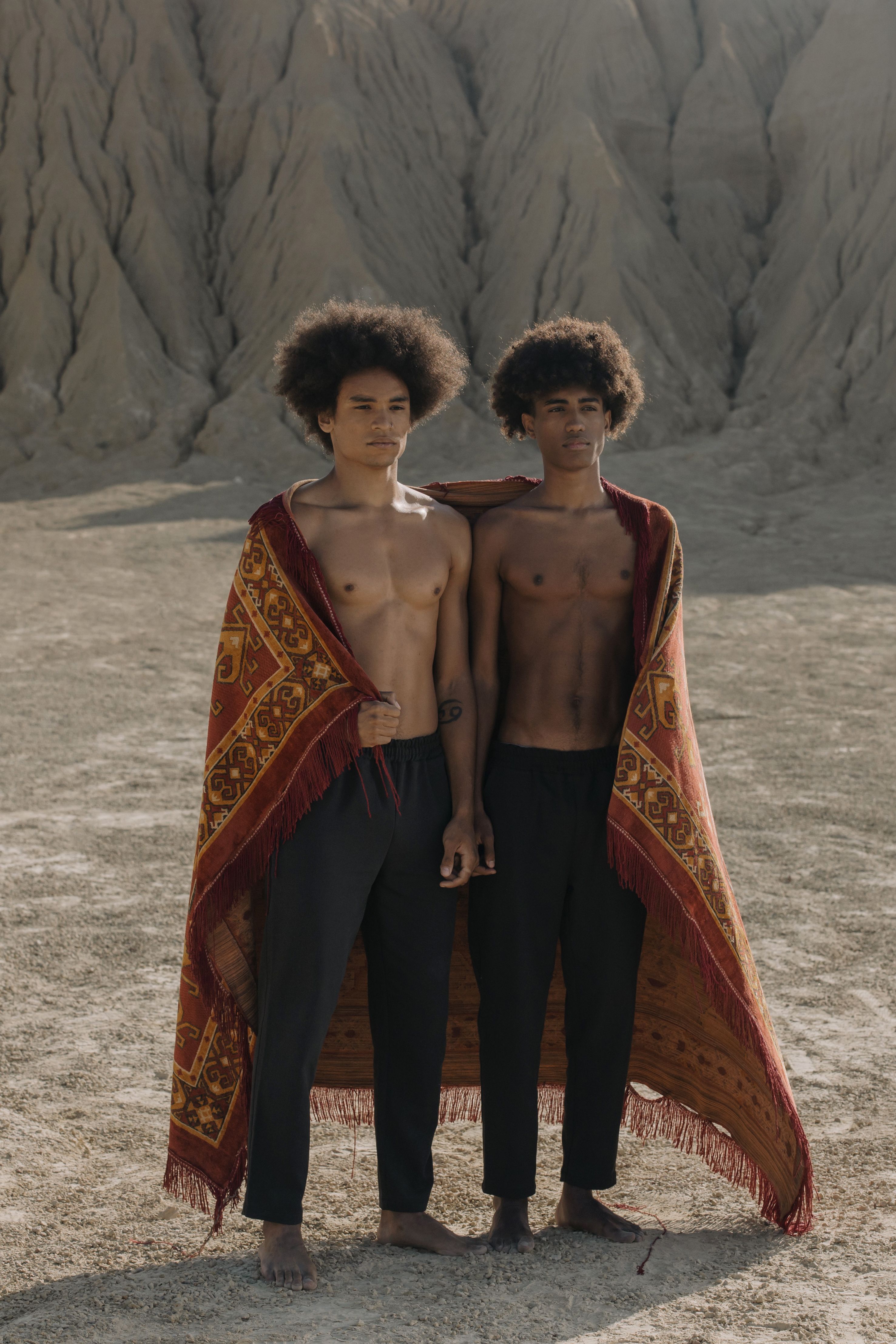 Two shirtless men with afro hairstyles stand in a desert landscape wrapped in a printed blanket.