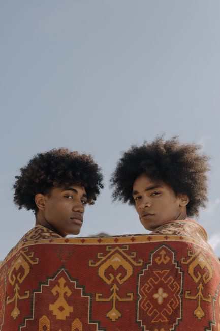 Two young men with afro hair wrapped in a patterned blanket under a clear blue sky.