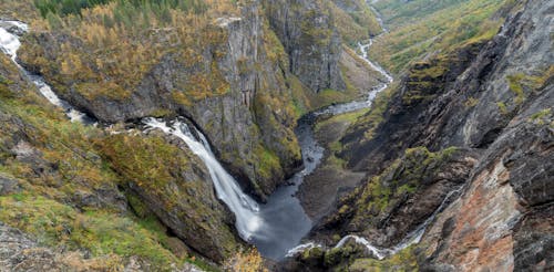 From above of rough rocky terrain covered with green moss and waterfall flowing into narrow river in highland in nature