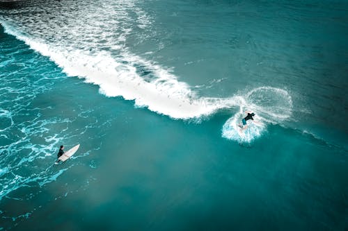 Aerial view of unrecognizable sporty surfers riding surfboard and wakesurfing on azure seawater and leaving foamy trail behind