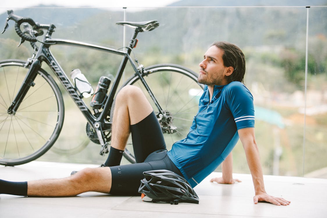 Free Side view of young contemplative fit male athlete in sportswear sitting near bicycle on fenced terrace while looking up Stock Photo