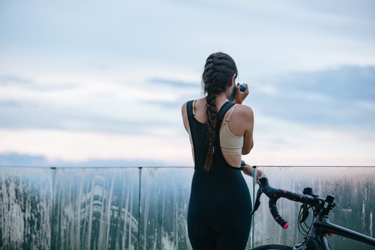Back view of unrecognizable sportswoman drinking beverage from mug near bicycle and fence while having break