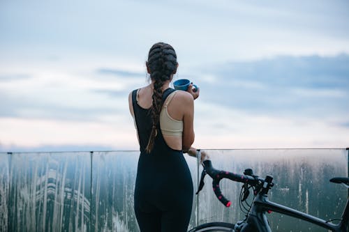 Free Back view of anonymous cyclist in sportswear with cup of beverage near fence and bike contemplating cloudy sky Stock Photo