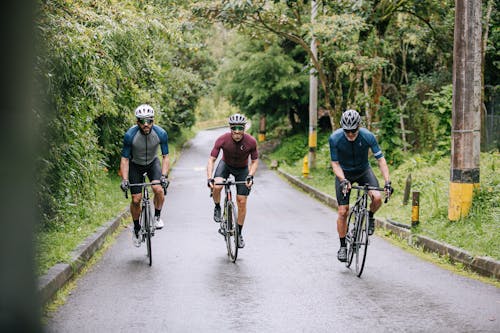 Unrecognizable male athletes in active wear and helmets riding bikes on asphalt roadway between lush green trees