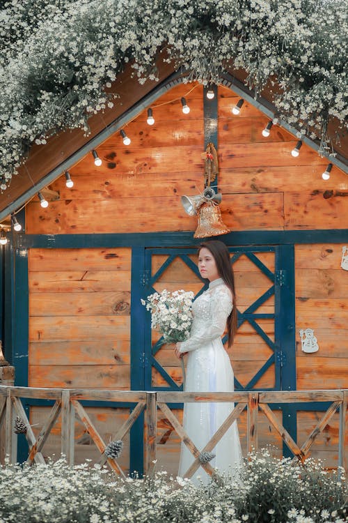 Woman in White Wedding Gown Holding Bouquet Standing Beside Brown Wooden House