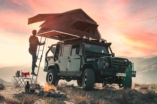 Back view of crop anonymous male traveler watching sunset during camping in mountains and climbing upstairs to tent located on roof of contemporary offroader