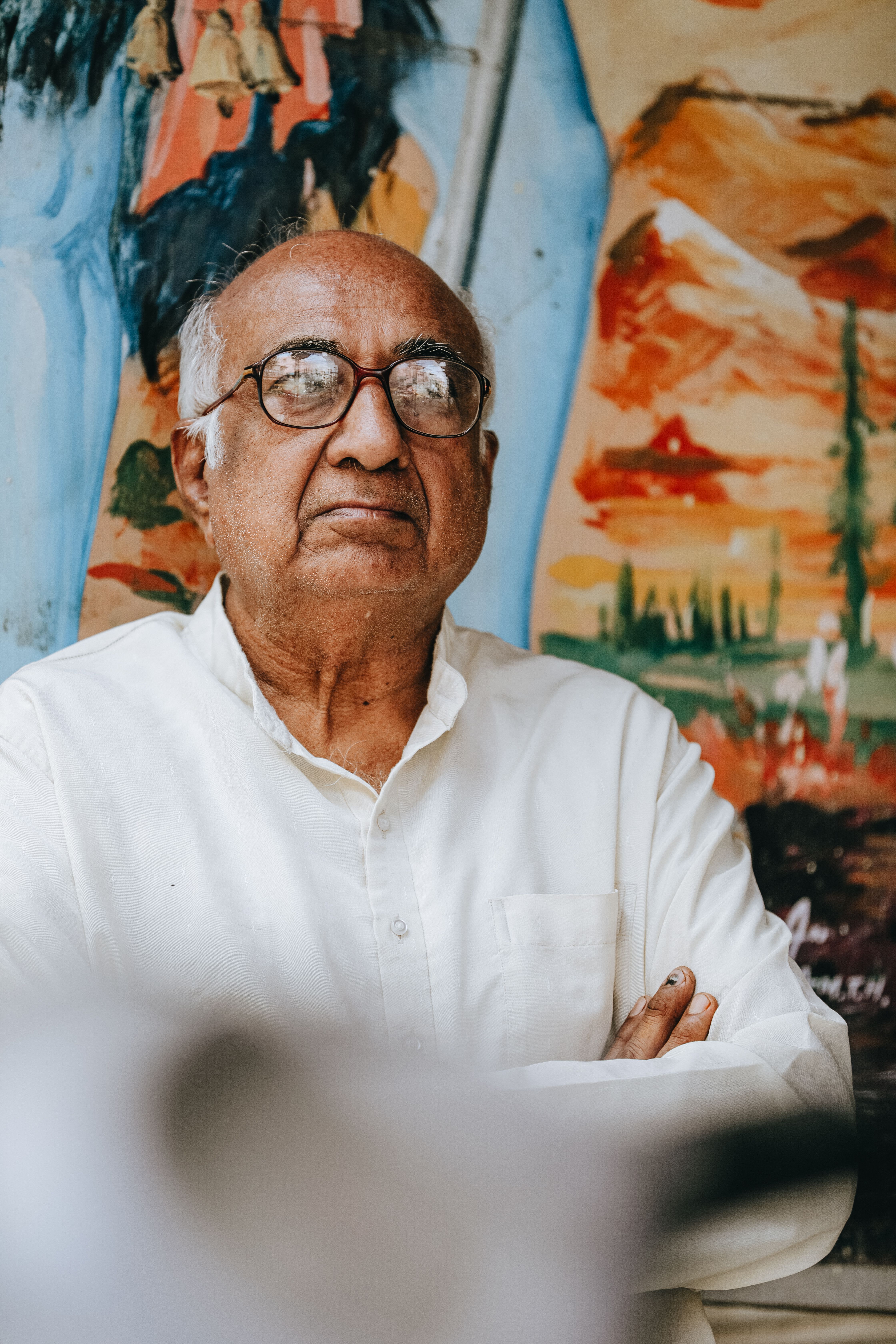 Elderly man with eyeglasses poses confidently in front of his vivid artwork.