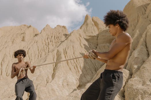 Two shirtless men engage in a competitive tug-of-war in a desert setting.